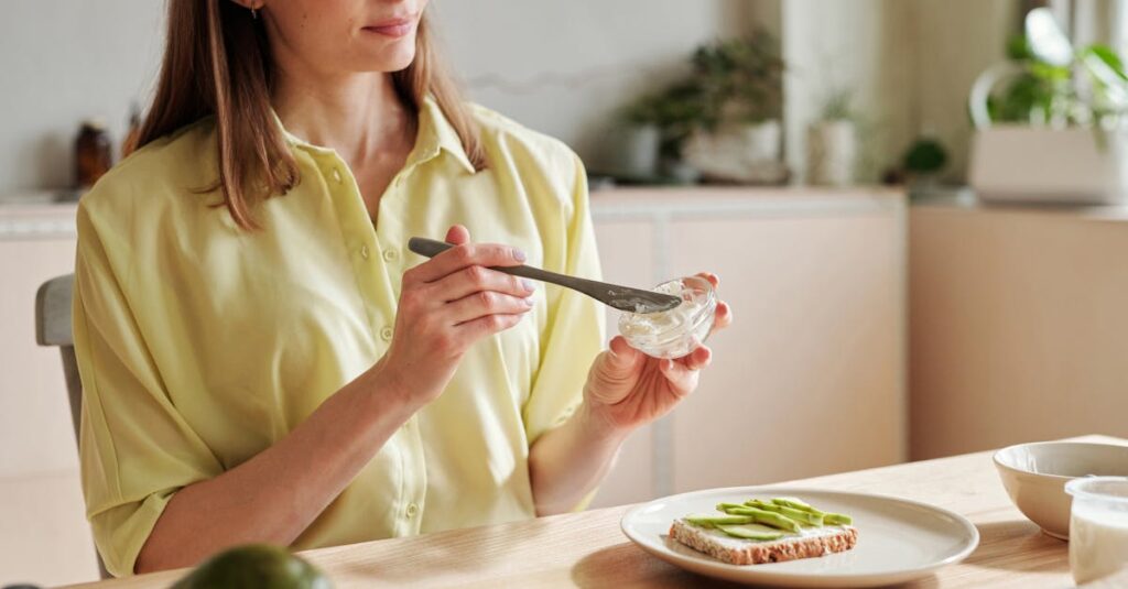 Young woman spreading cream cheese on toast with sliced avocado, creating a healthy breakfast.
